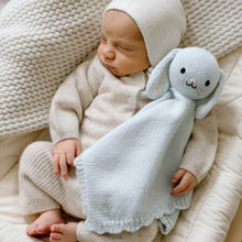 Load image into Gallery viewer, A sleeping newborn baby wearing a cream knit onesie and matching hat, holding a pale blue knitted bunny comfort blanket in a soft cream crib.