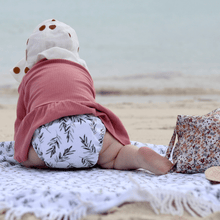 Load image into Gallery viewer, Baby sitting on a blanket at the beach wearing a pink top and leaf-patterned cloth nappy.