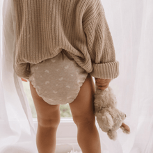 Load image into Gallery viewer, Toddler wearing a beige knit sweater and patterned beige cloth nappy holding a teddy bear in a softly lit room.