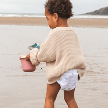 Load image into Gallery viewer, Child in a beige sweater and cloth nappy standing on a beach holding a pink bucket.