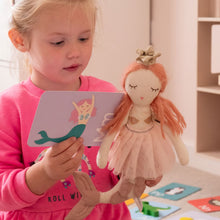 Load image into Gallery viewer, Child holding a doll and a book with a mermaid illustration in a room setting.
