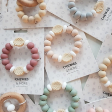 Load image into Gallery viewer, A styled flat-lay of multiple silicone teething chew rings on branded cards. The set includes pastel pink, peach, mint green, and light blue beads with natural wood leaf accents, alongside a wooden ring teether.
