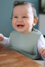 Load image into Gallery viewer, Baby wearing a light green silicone bib with front pocket sitting at a table with a blurred background

