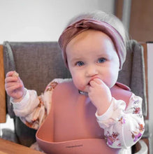 Load image into Gallery viewer, Baby wearing a pink silicone bib with front pocket and headband with a bow, sitting in a high chair.
