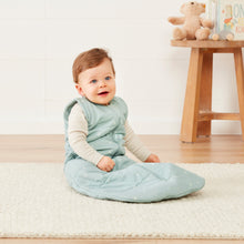 Load image into Gallery viewer, Baby in a light blue sleeping bag sitting on a white rug with a wooden stool and teddy bear in the background.
