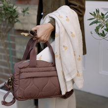 Load image into Gallery viewer, Person holding a brown quilted changing bag with a pears design muslin draped over their arm, standing outdoors.
