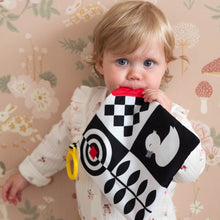Load image into Gallery viewer, Child holding a black and white checkered sensory square with a red circle and yellow handle against a floral-patterned wall.
