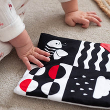 Load image into Gallery viewer, Baby's hand reaching towards a black fabric with white and red patterns on a beige carpet.
