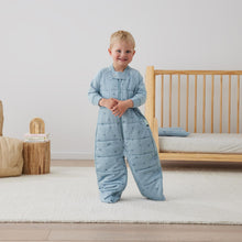 Load image into Gallery viewer, Child wearing a blue quilted ErgoPouch sleepsuit standing in a bedroom with a wooden crib and neutral decor.
