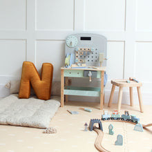 Load image into Gallery viewer, Children's play area with wooden toys, a clock, and a stool against a white paneled wall.
