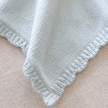 Load image into Gallery viewer, A close-up of a pale blue knit blanket with a fringed edge, laid over a soft beige fabric background.
