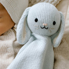 Load image into Gallery viewer, A close-up of a knitted bunny in light blue, shown resting on a baby's chest. The soft toy has long floppy ears, embroidered black eyes and mouth, and a tiny pink nose.
