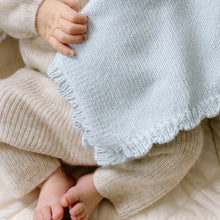 Load image into Gallery viewer, A close-up lifestyle shot of a baby wearing a cream ribbed knit outfit and holding a pale blue knit blanket with a scalloped edge.

