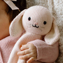 Load image into Gallery viewer, A close-up of a person holding a knitted bunny comforter in a soft pink sweater. The bunny has long floppy ears, black embroidered eyes and mouth, and a small pink nose, set against a cozy white textured blanket background.
