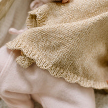 Load image into Gallery viewer, Close-up of a knitted baby blanket in a warm beige color with a ruffled edge, draped over light pink clothing.
